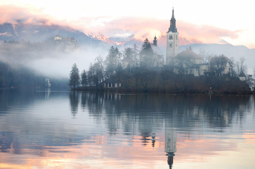 The Bled island church reflecting in the water with the snowy mountains and the Bled castle in the background