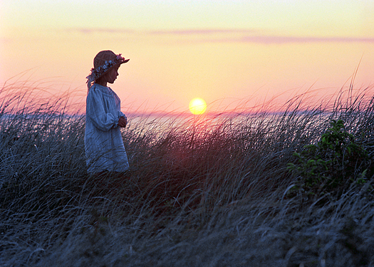 A young girl in a sunhat walking through grass with sunset over the ocean as a backdrop