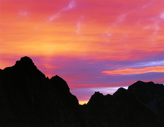Silhouetted mountain landscape against pink-orange sunset by John Fielder